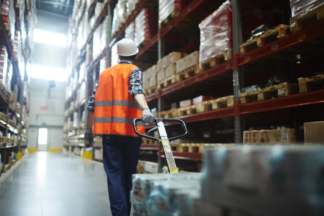 A worker moving a pallet down a warehouse aisle towards the outbound dock